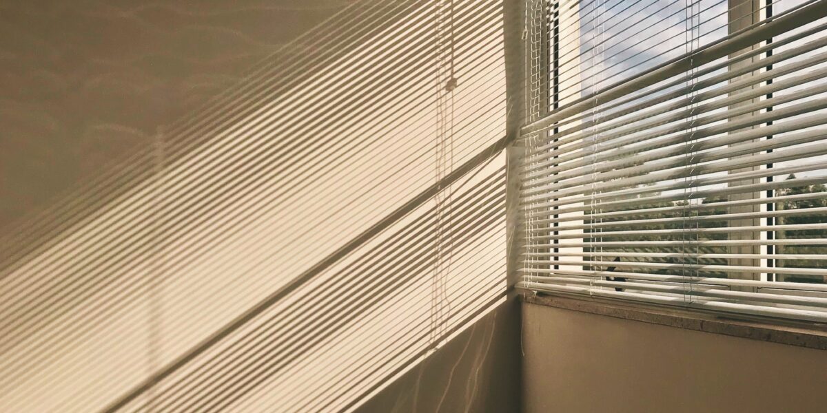 Lys skinner inn gjennom halvåpne persienner i et vindu. Den beige veggen lyses delvis opp, rommet ser tomt ut.