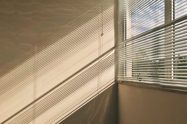 Lys skinner inn gjennom halvåpne persienner i et vindu. Den beige veggen lyses delvis opp, rommet ser tomt ut.